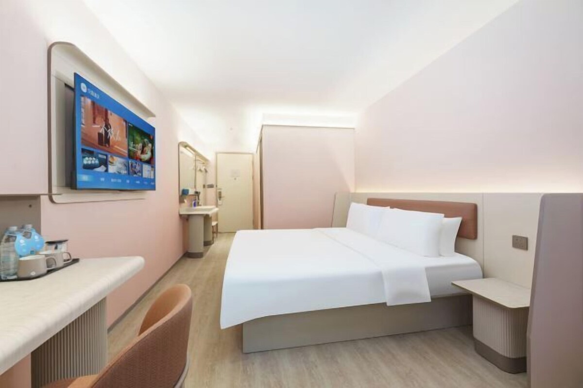Quarto de hotel com cama, escrivaninha, cadeira, televisão na parede e banheiro visível por uma porta aberta.