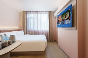 Quarto de hotel com cama, televisão na parede, escrivaninha com cafeteira e janela com cortinas.