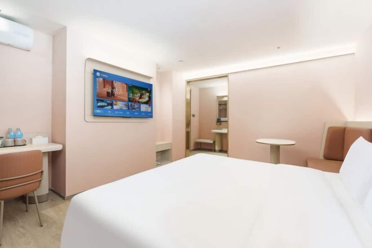 A hotel room with a bed, a desk, a chair, a small table, and a wall-mounted screen.