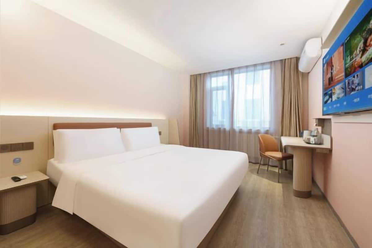 A hotel room with a large bed, a desk with a chair, a small table, and a wall-mounted TV.