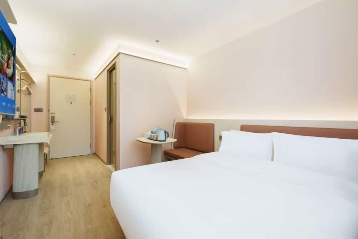 A hotel room with a large bed, a small bedside table, a chair, and a door leading to another room.