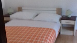 Een bed met een geruit dekbed, een wit matras en twee kussens. Er staat een nachtkastje met een lamp en een lade.