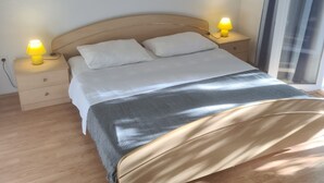 Een bed met twee witte kussens en een grijze deken, omringd door twee nachtkastjes met gele lampen, in een kamer met houten vloeren en een groot raam.