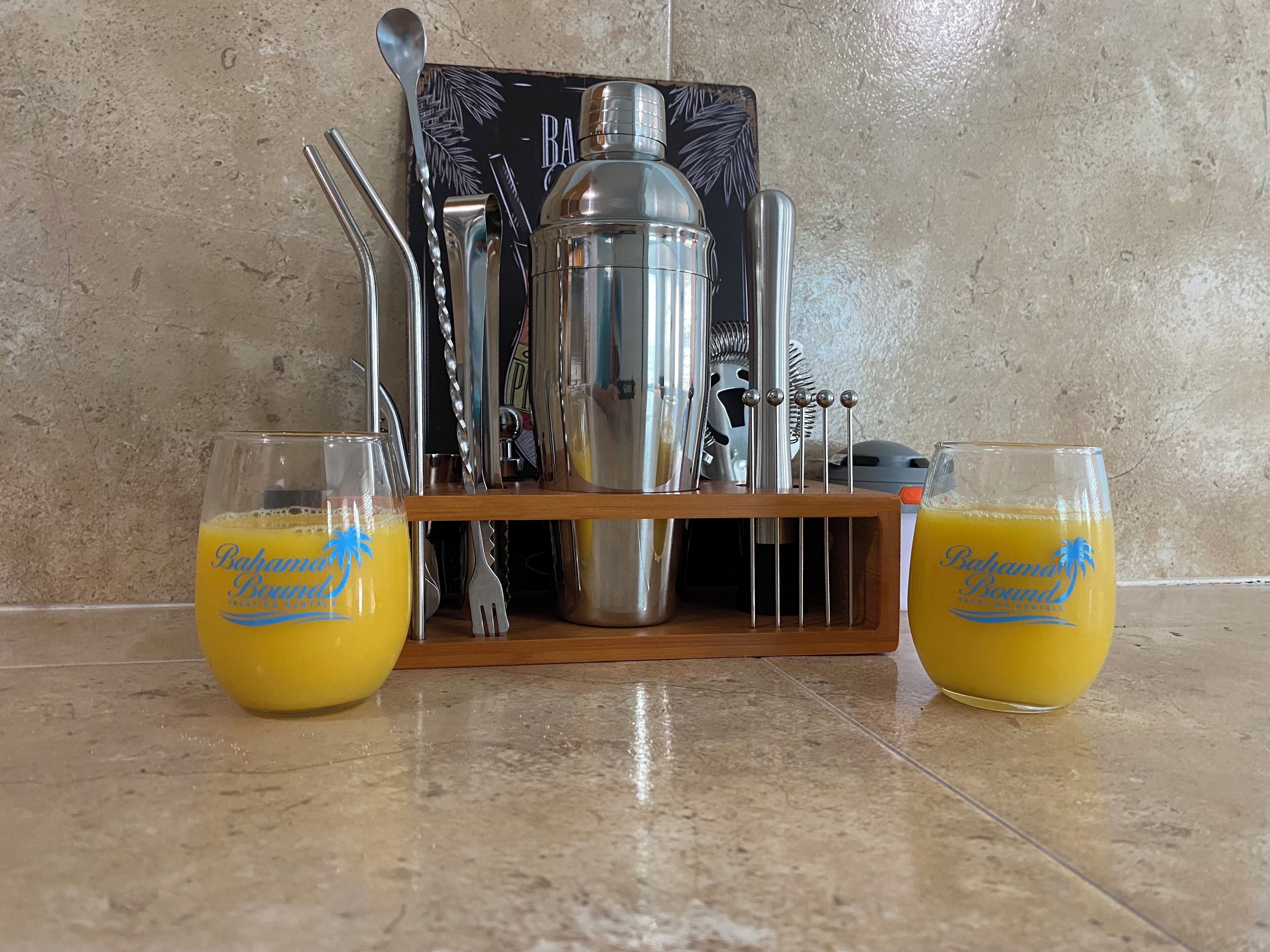 Due bicchieri di succo d'arancia su un pavimento piastrellato, con un set da bar che include uno shaker, un misurino e altri accessori.