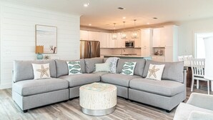 A modern living room with a sectional sofa, a round coffee table, and a kitchen in the background. - Beachfront House with Pool! 6 BR/6.5 Beachfront Paradise! (Gulf Shores)
