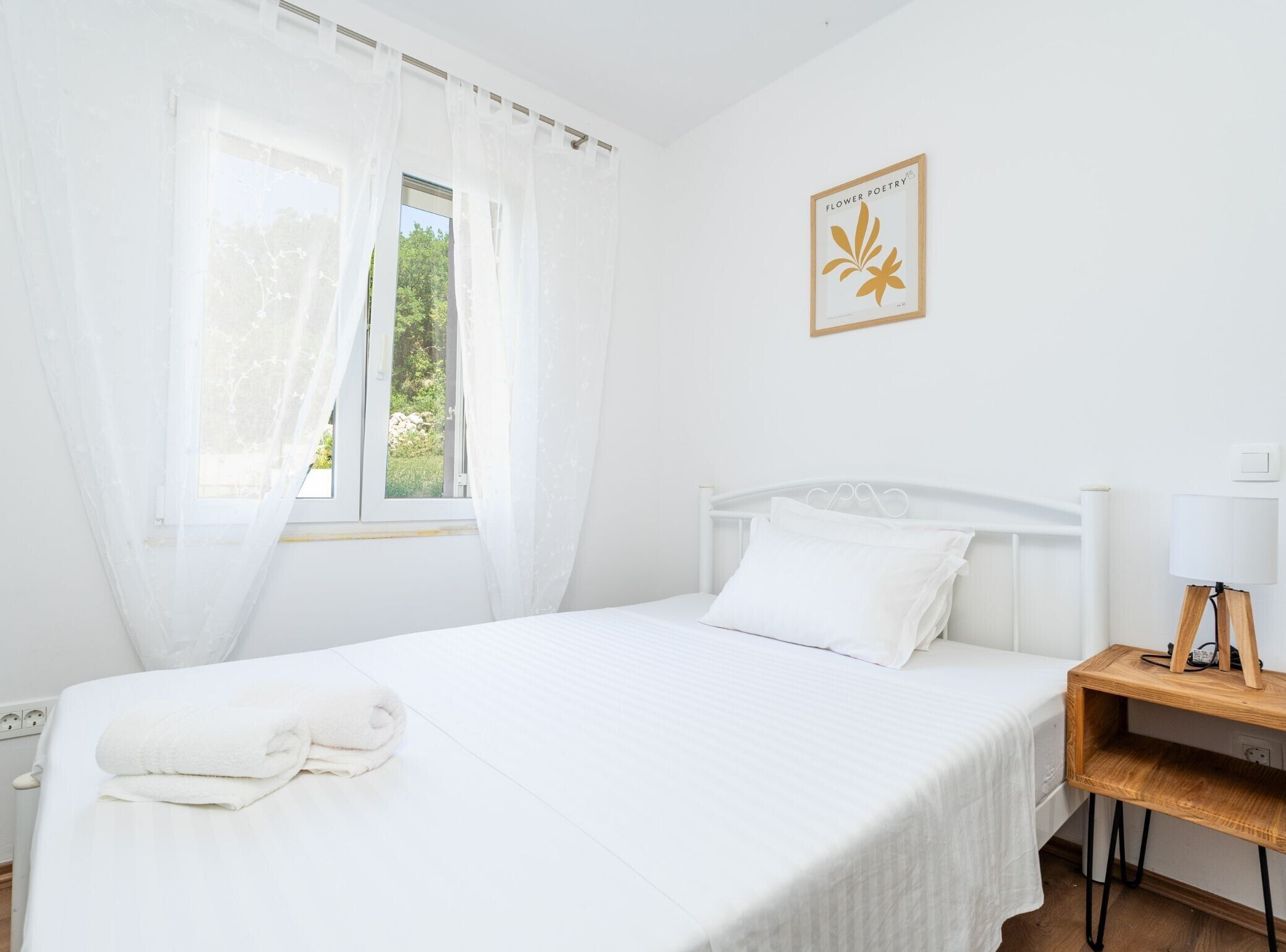 Une chambre à coucher propre et blanche, avec un lit, une table de chevet dotée d’une lampe et une fenêtre ornée de rideaux légers.