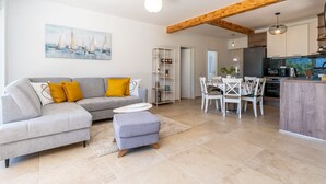 A modern living room with a grey sofa, ottoman, and dining table with chairs. There is a kitchen area with a refrigerator, microwave, and wooden beams on the ceiling.