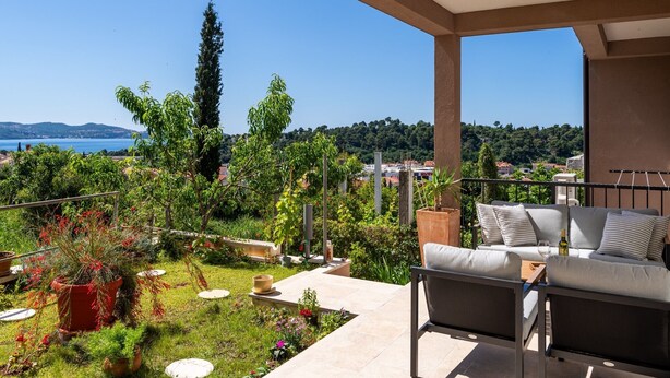 A balcony with a sofa and table, overlooking a garden with various plants and a view of the sea and hills.