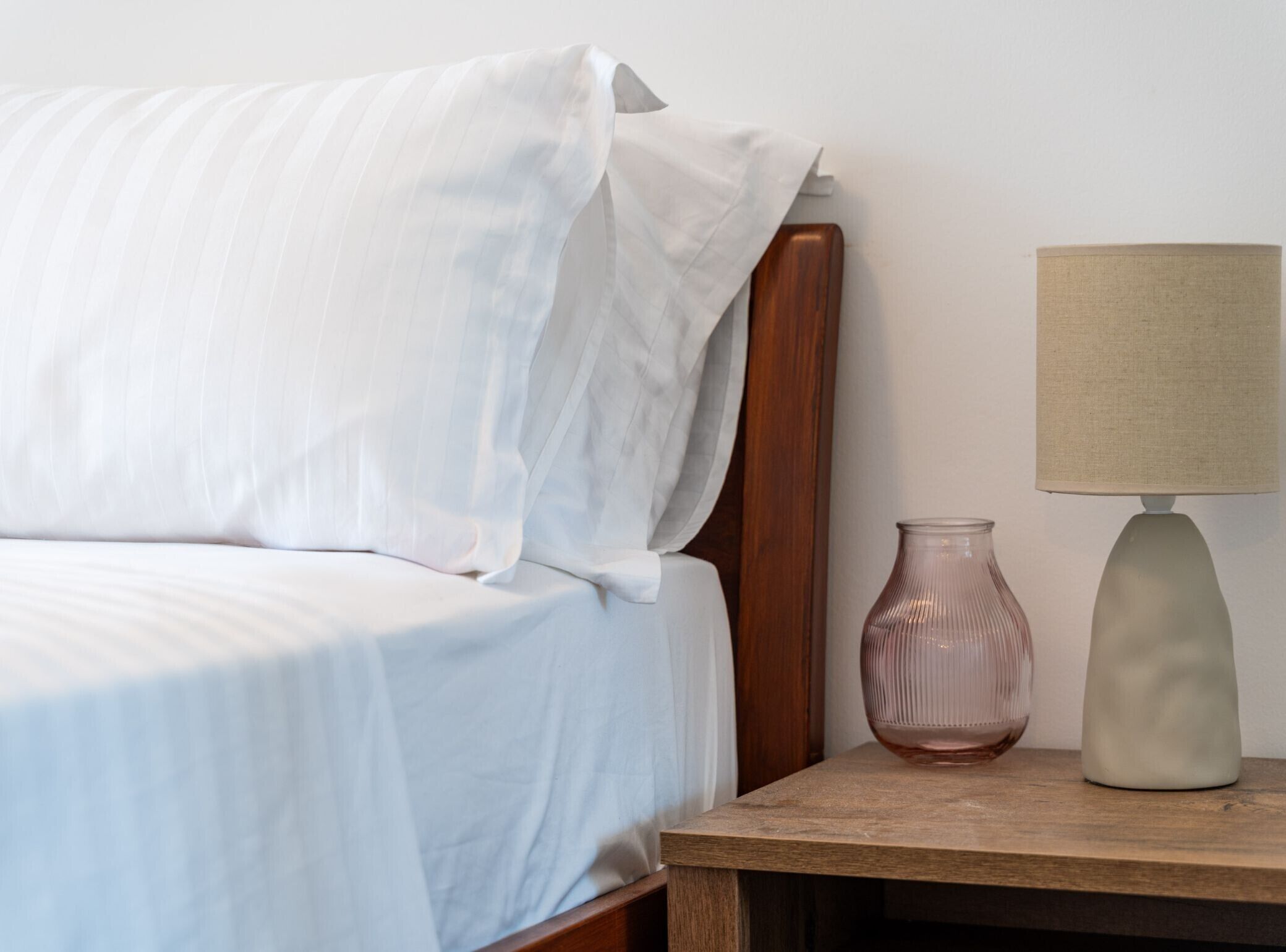 Un lit bien fait, avec du linge de lit blanc, une table de chevet en bois avec une lampe et un vase, et une tête de lit en bois.