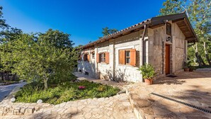 Una casa de piedra con techo de tejas, contraventanas de madera y un pequeño jardín con plantas en macetas.