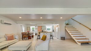 A spacious living area with a sofa, ottoman, and a dining table with chairs. A kitchen with a central island and a staircase leading to an upper level is visible. - Charming Catskills Getaway: Hike, Bike, Sip & More (Acra)