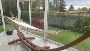 A hammock set up inside a room with large windows, offering a view of a garden and a red-roofed building outside. - Ferienhaus in Herzwolde für 4+2 Personen (Wokuhl-Dabelow)