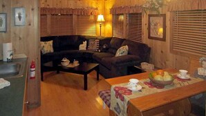 Ein gemütliches Wohnzimmer mit einer Sofagarnitur, einem Couchtisch und einem Esstisch mit Obstschale. Holzverkleidung an den Wänden und eine Decke mit freiliegenden Balken.