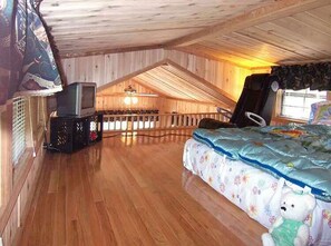 Une chambre confortable située dans les combles, avec un plancher en bois, un lit recouvert d’une couette à motifs floraux, une télévision et une fenêtre offrant une belle vue.