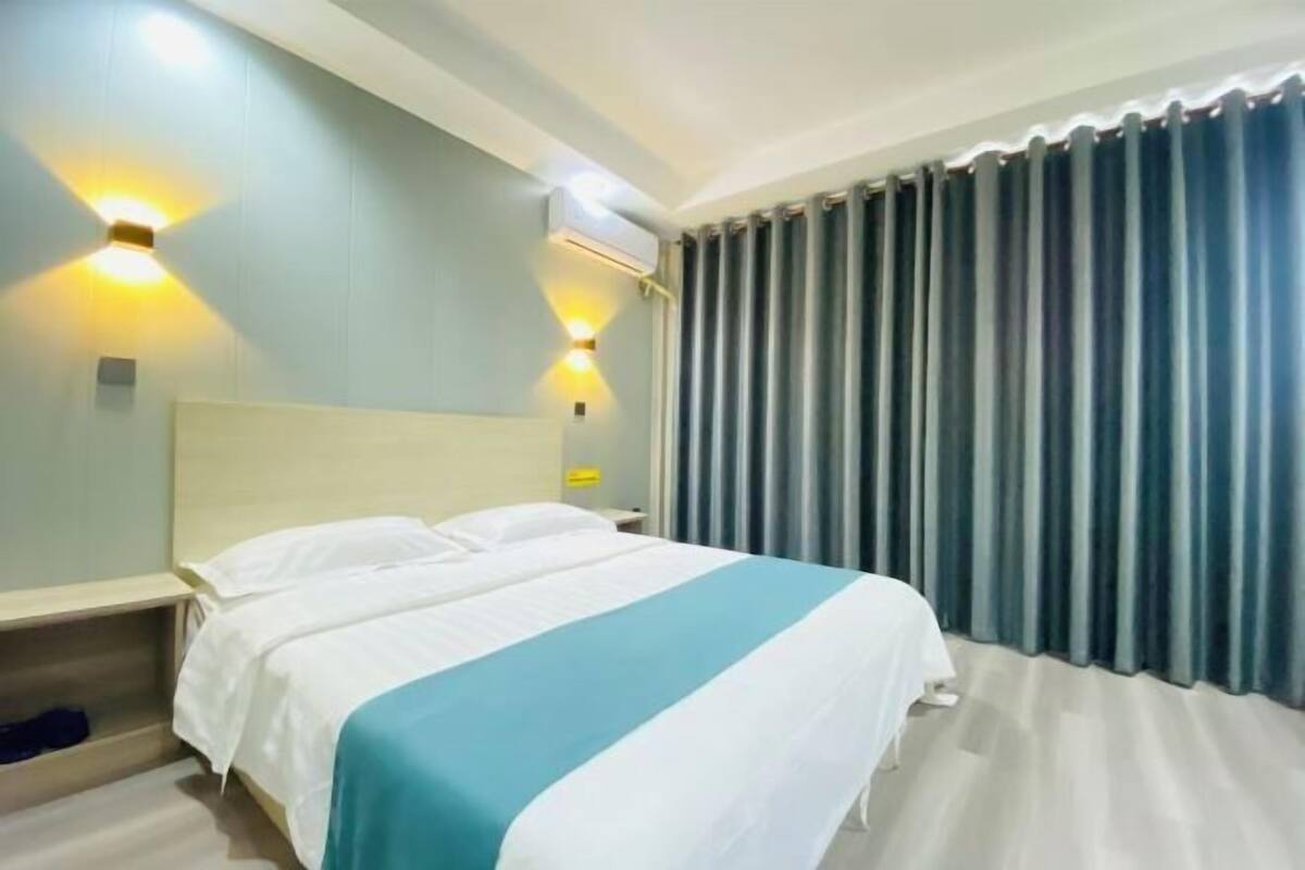 A hotel room with a bed, a nightstand, a wall-mounted air conditioner, and a window with curtains.