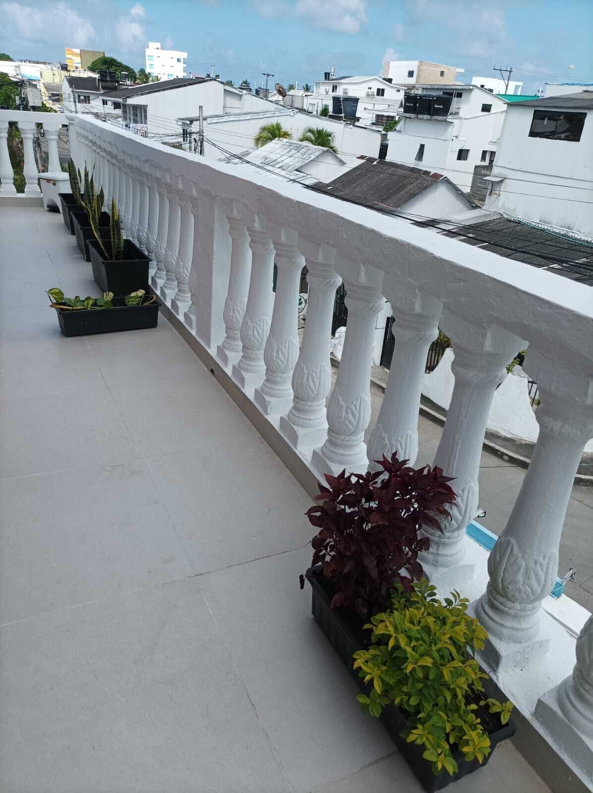 Un balcón con barandillas blancas, plantas en macetas y vistas de edificios circundantes.