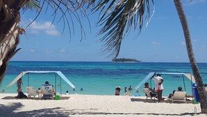 A beach with clear turquoise waters, a small island in the distance, and people relaxing under shaded areas. - HOTEL VERANO AZUL (San Andrés)