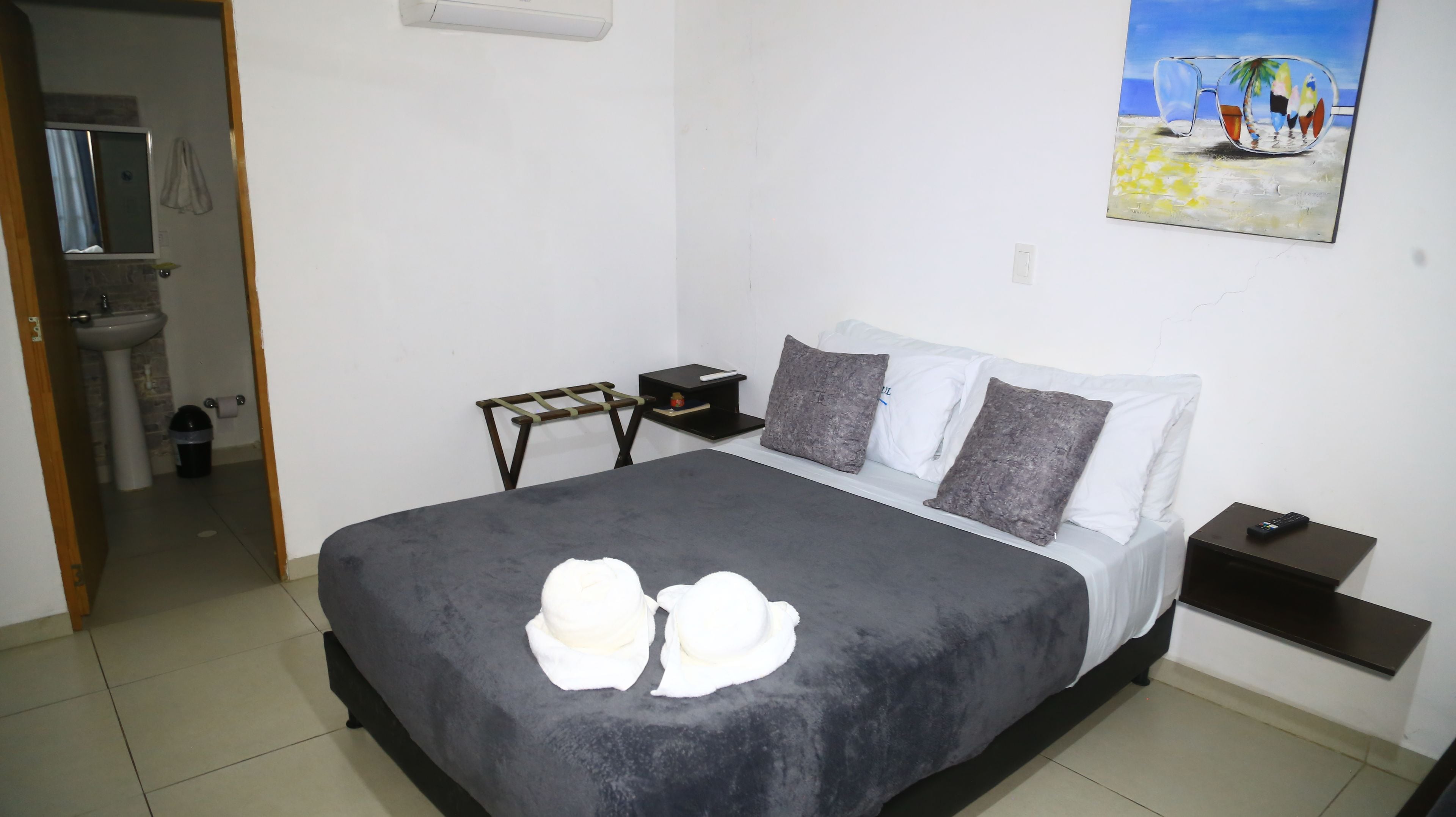 Un dormitorio con cama, mesita de noche, una pequeña mesa, un cuadro y un baño visible a través de una puerta abierta.