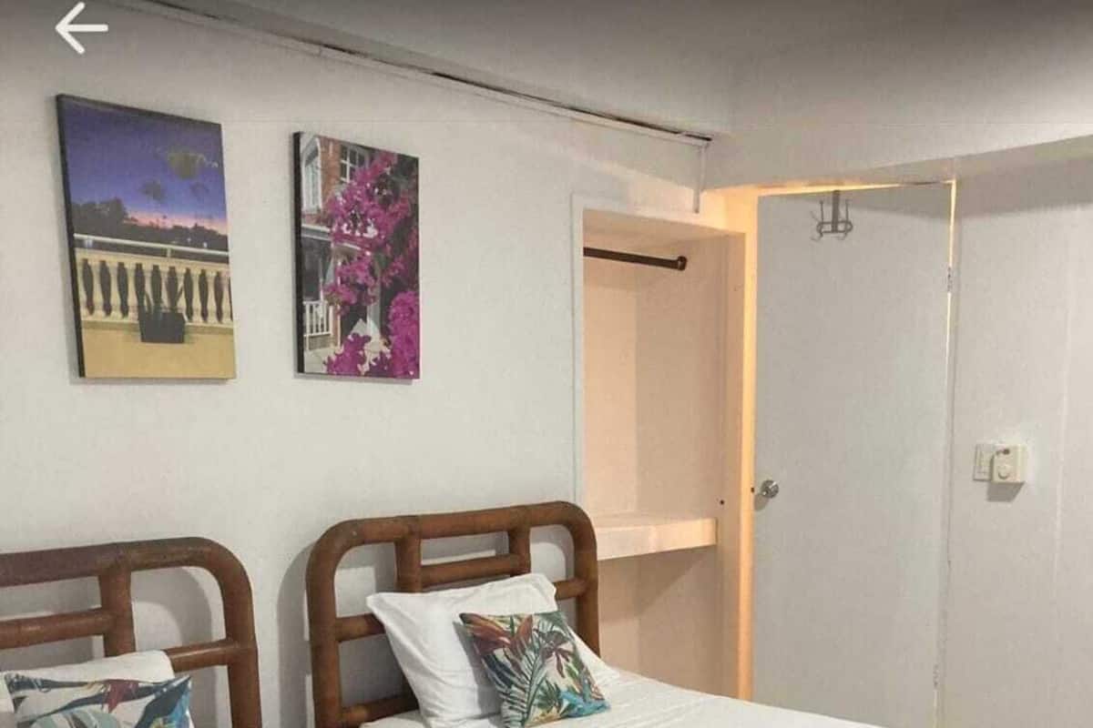 Quarto com duas camas de solteiro, cada uma com cabeceira de madeira e roupa de cama branca. Há duas pinturas na parede, uma retratando um pôr do sol e outra com flores cor-de-rosa. Uma porta é visível ao fundo.