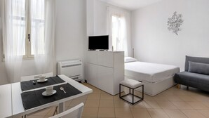 A compact room with a bed, a small table with a cup and spoon, a TV on a cabinet, and a gray sofa. - White Leaves Studio by Wonderful Italy (Bologna)