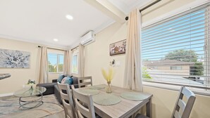 Un salon moderne avec un coin repas, comprenant un canapé, une table basse en verre, une table à manger en bois avec des sets de table, et une grande fenêtre avec des stores.
