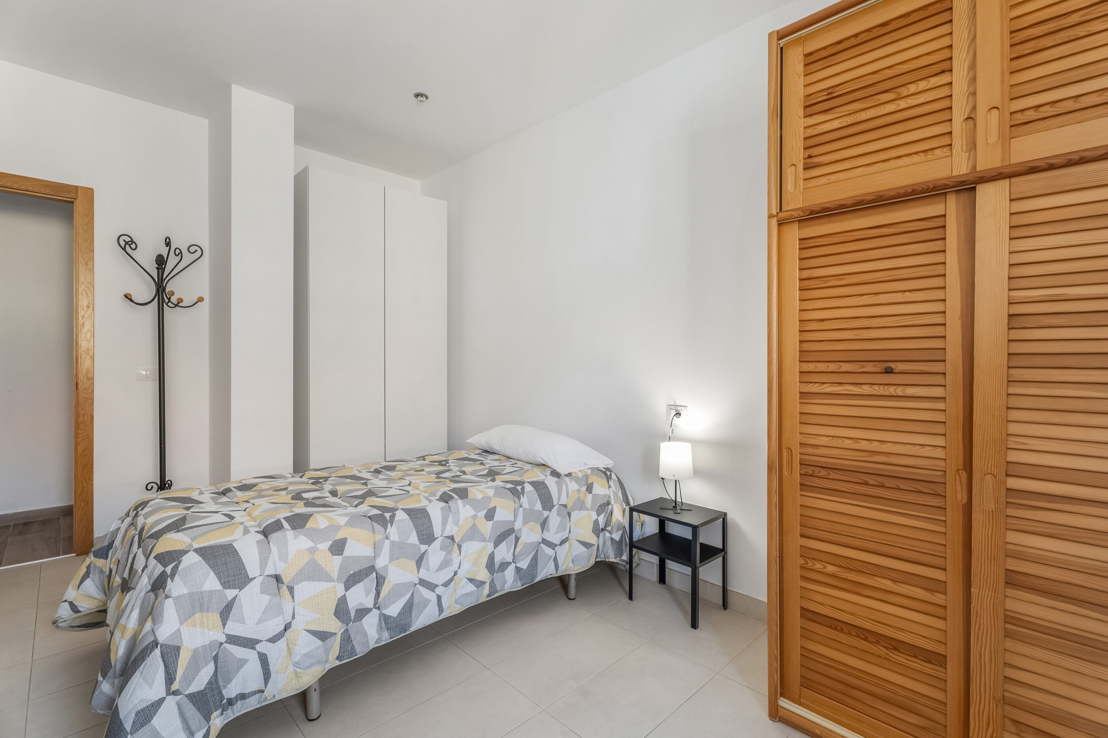 A bedroom with a bed, a nightstand with a lamp, a coat rack, and a wooden wardrobe.