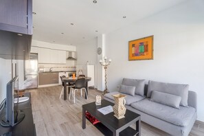A modern living room with a grey sofa, a coffee table, and a TV. There is a dining area with a table and chairs, and a kitchen in the background.