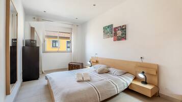 A modern bedroom with a bed, bedside tables, a nightstand, a suitcase, and artwork on the wall.