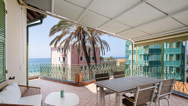 A balcony with a table and chairs, a view of the sea, and a building with a pink facade.