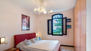 A bedroom with a red bed, a window with a view of greenery, a white radiator, and a wooden nightstand with drawers.