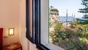 A room with a window overlooking a coastal landscape, a bedside table with a lamp, and a wall-mounted light.