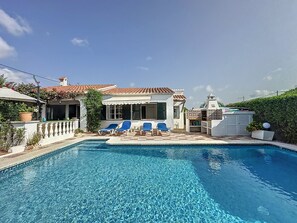 A swimming pool with blue water, surrounded by a white building with a tiled roof, and blue lounge chairs.