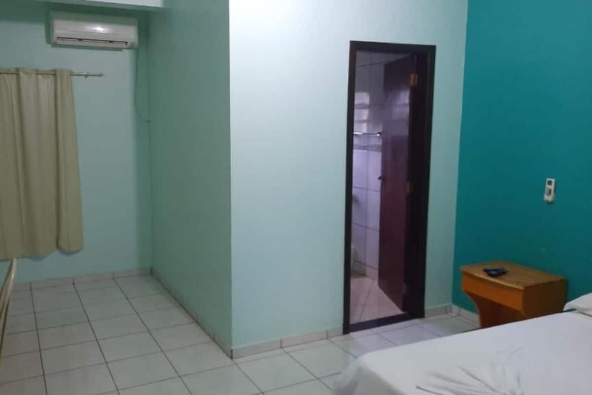 Una habitación con una cama, una mesita de noche de madera, una cortina y una puerta que da a otra habitación.
