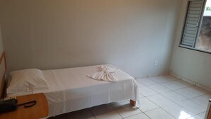 Ein kleines, sauberes Zimmer mit einem Einzelbett, einem Nachttisch aus Holz und einem Fenster mit Jalousien.