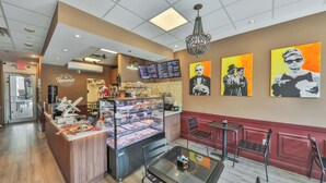 A bakery with a display case of pastries, a coffee machine, and a menu board. There are chairs and tables for seating, and a chandelier hangs from the ceiling.