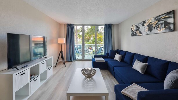 A modern living room with a flat-screen TV, a sofa, a coffee table, and a balcony with a view of palm trees.