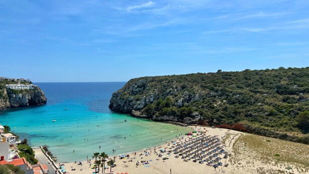 Une plage côtière bordée d’une falaise rocheuse, avec une eau d’un bleu limpide et une rangée de parasols de plage.