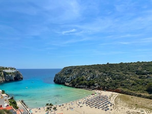 A coastal beach with a rocky cliff, clear blue water, and a row of beach umbrellas. - Apartment 'Ca Nostra' with Balcony and Wi-Fi (Cala en Porter Menorca)