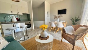 Une cuisine moderne avec des meubles blancs, un coin repas avec une table et des chaises blanches, un fauteuil en osier et une table ronde agrémentée d’un vase de fleurs jaunes.