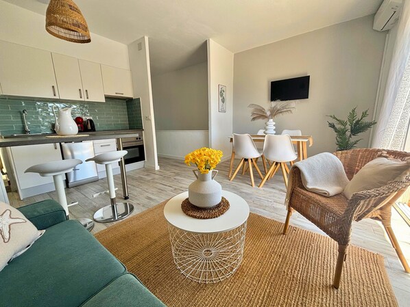 Una cocina moderna con armarios blancos, un comedor con mesa y sillas blancas, una silla de mimbre y una mesa redonda con un jarrón de flores amarillas.