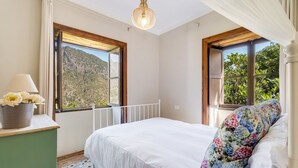 A bedroom with a bed, a nightstand, a lamp, a floral pillow, and a view of a mountainous landscape through large windows. - Holiday Home 'Caldera De Taburiente Rural' with Mountain View, Private Terrace and Wi-Fi (El Paso)