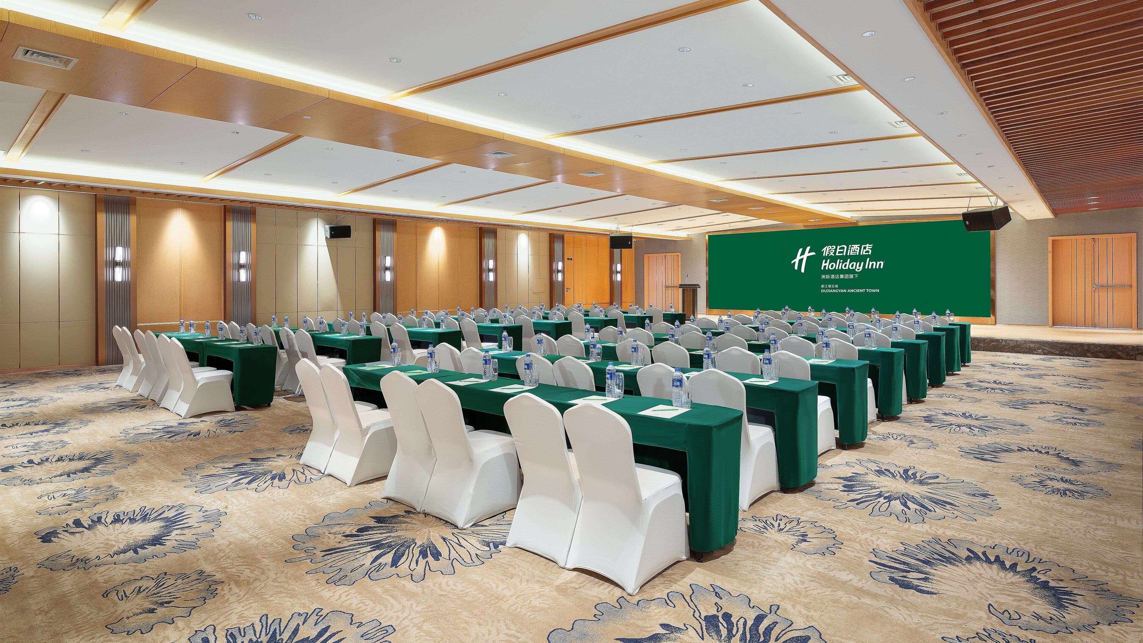 A conference room with rows of tables and chairs, a green carpet, and a projection screen in the background.