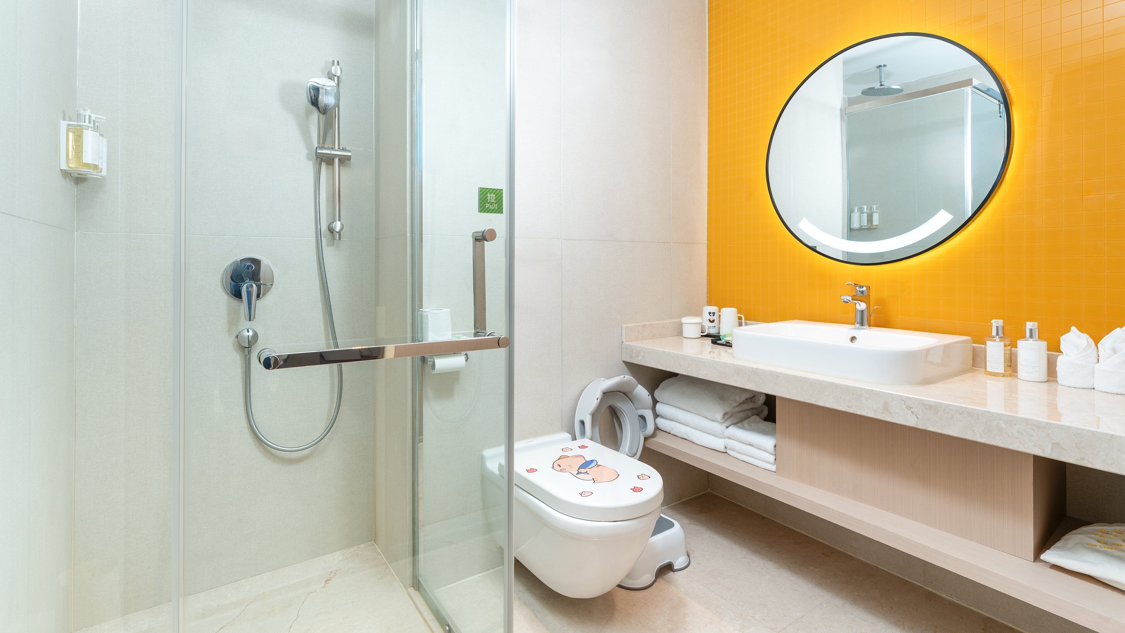 Ein modernes Badezimmer mit einer gläsernen Duschkabine, einer weißen Toilette inklusive Kindersitz, einem Waschbecken mit Spiegel und einer gelb gefliesten Wand.