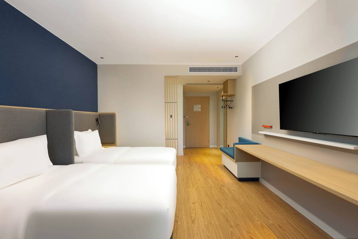 A hotel room with a large bed, a TV, a wooden floor, and a hallway leading to a bathroom.