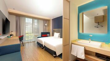 A hotel room with two beds, a TV, a desk, a chair, a large window with curtains, a bathroom with a sink and mirror, and a towel hanging on the wall.