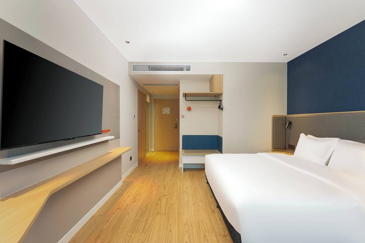 A modern hotel room with a large flat-screen TV, a bed with white linens, a wooden floor, and a blue accent wall.