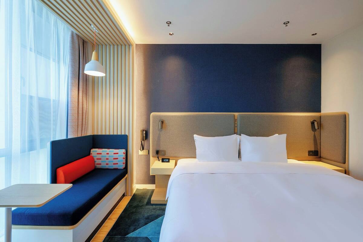 A hotel room with a bed, a sofa with a red cushion, a bedside table, and a nightstand with a lamp.