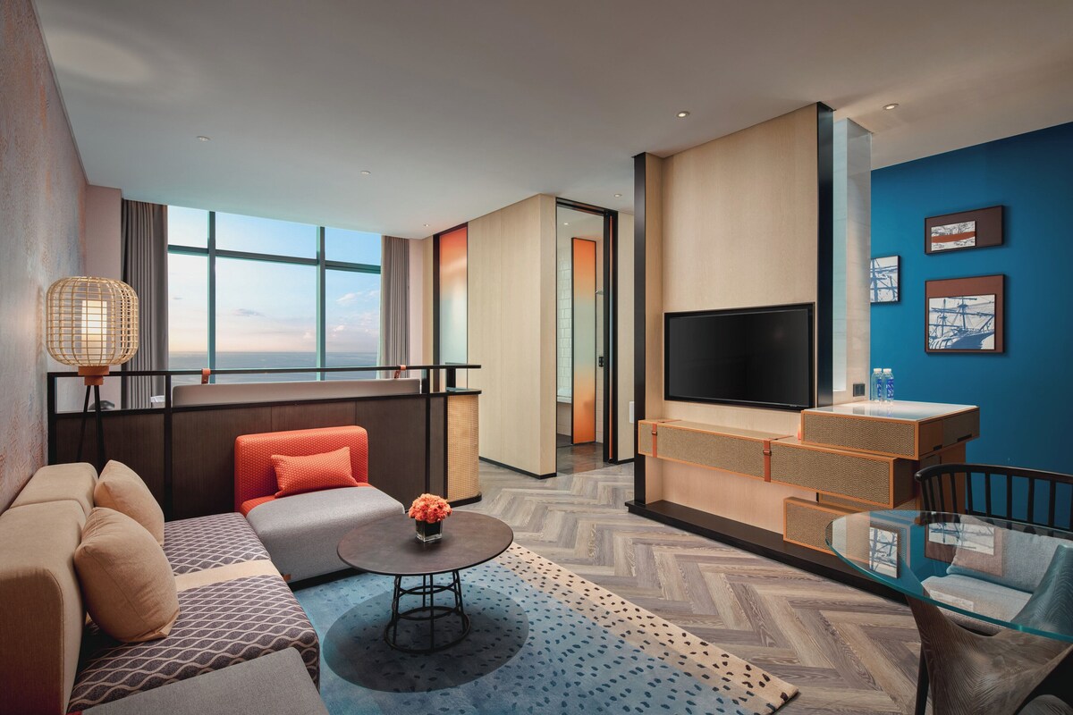 Une chambre d’hôtel moderne équipée d’un canapé, d’une table basse, d’une télévision et d’une grande fenêtre donnant sur la ville.