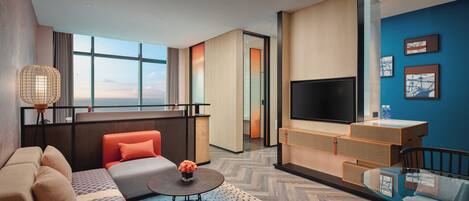 A modern hotel room with a sofa, a coffee table, a TV, and a large window offering a city view.