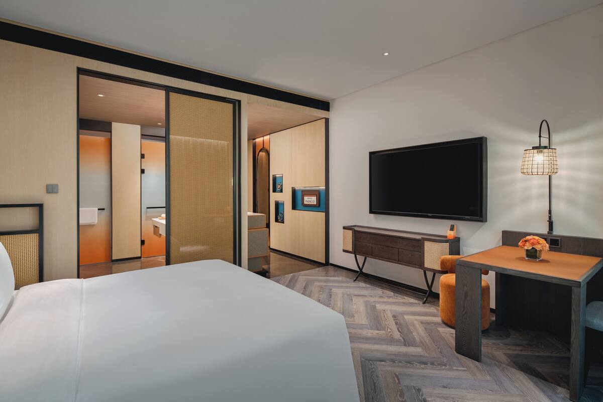 Een moderne hotelkamer met een groot bed, een bureau met een flatscreen televisie, een lamp en uitzicht op de badkamer en gang.